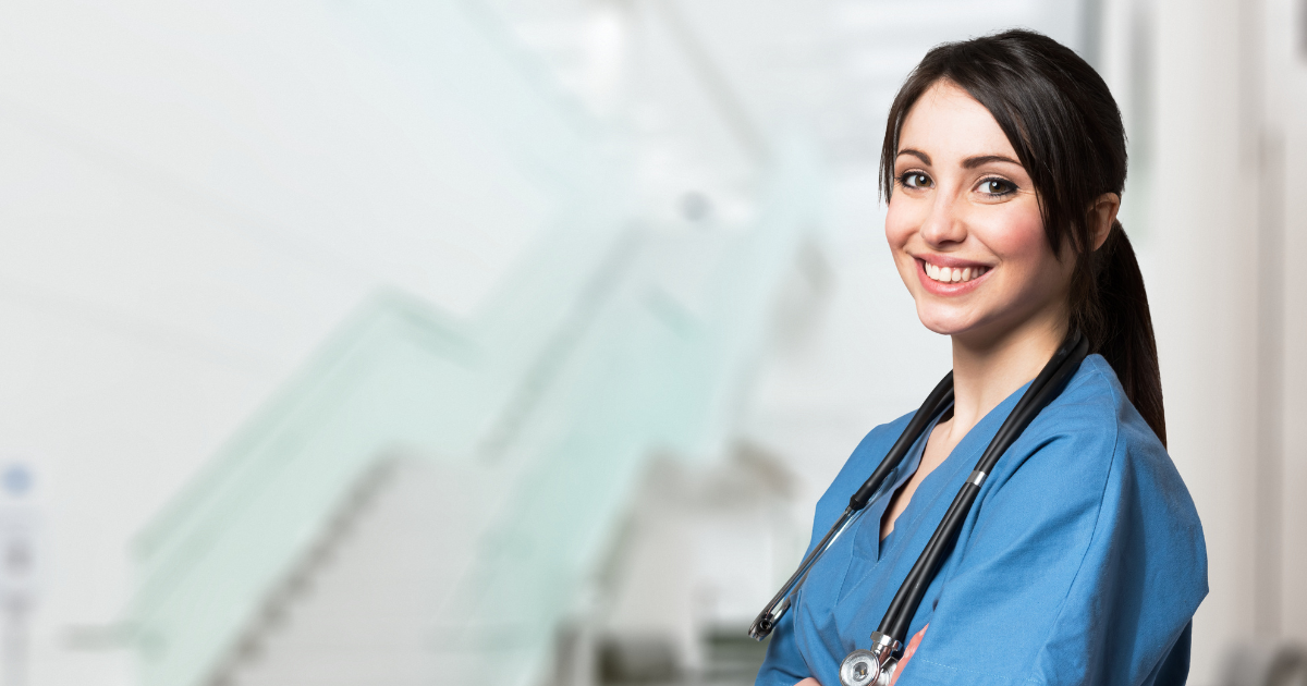 Female caregiver in blue scrubs stands with arms crossed and stethoscope around her neck with a big simile on face in front of staircase in a home care agency.