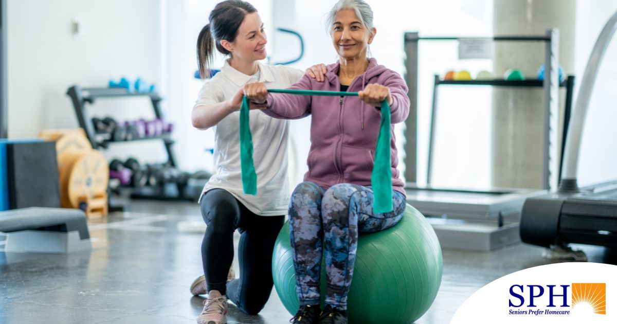 A care provider helps an older woman exercise with a resistance band and an exercise ball, representing how exercise can help with senior fall prevention.