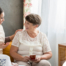 Smiling caregiver with her hand on an older woman’s shoulder as they share tea on the couch, showing home care vs assisted living