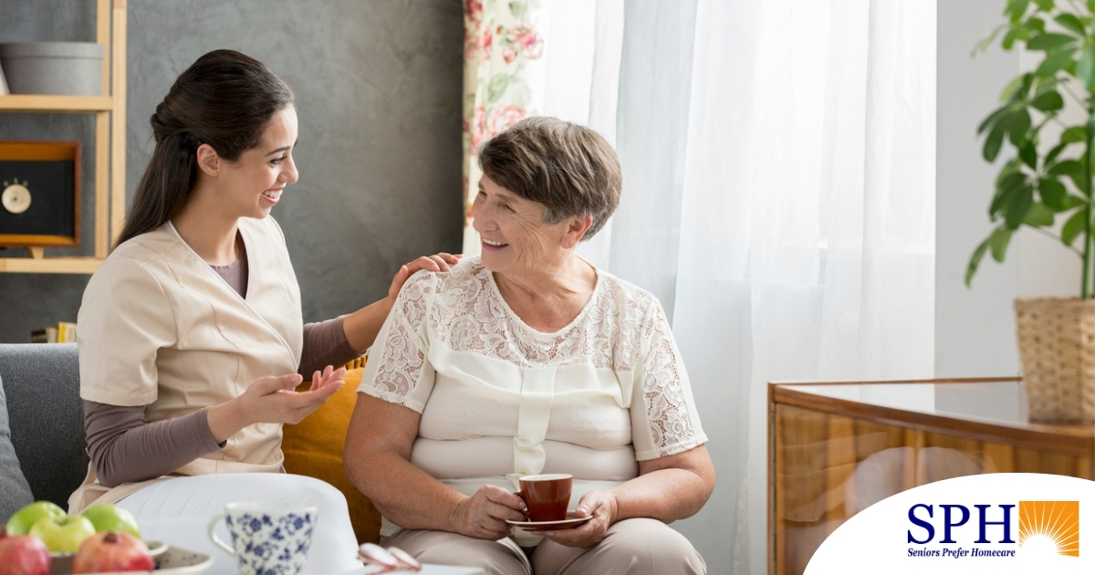 Smiling caregiver with her hand on an older woman’s shoulder as they share tea on the couch, showing home care vs assisted living